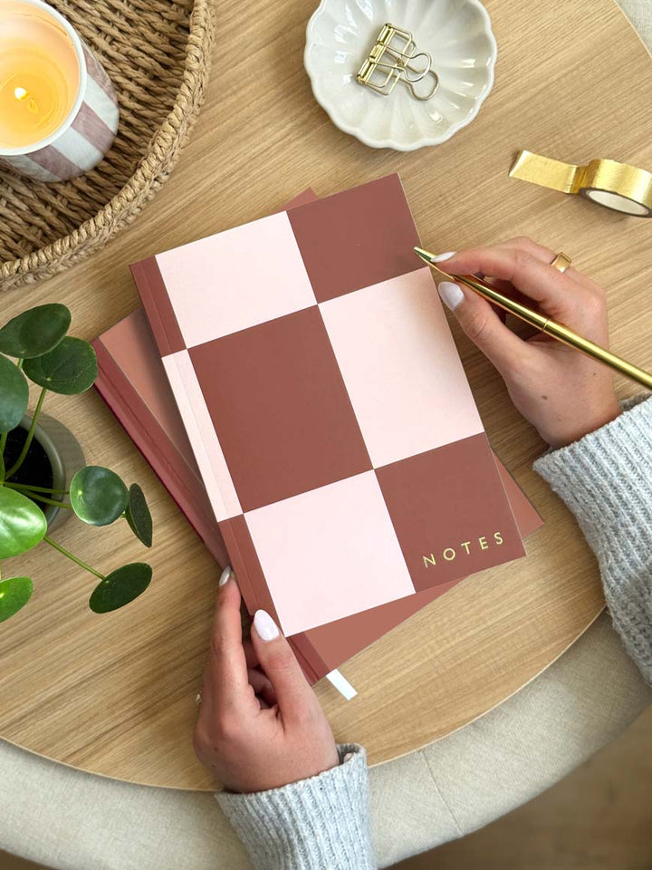 Person holding a checkered notebook with 'NOTES' on a wooden table with a candle and plant.