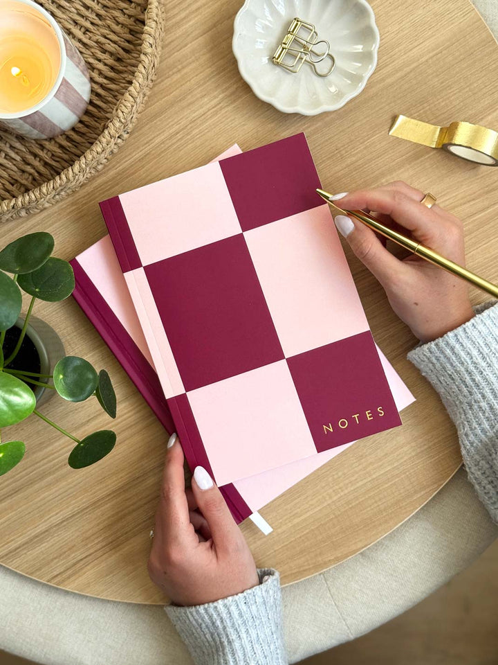 Woman holding a pink and red checkered notebook with 'NOTES' on a wooden desk.