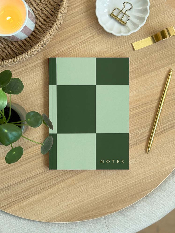 Green checkered notebook labeled 'NOTES' on a wooden desk with a candle and plant.