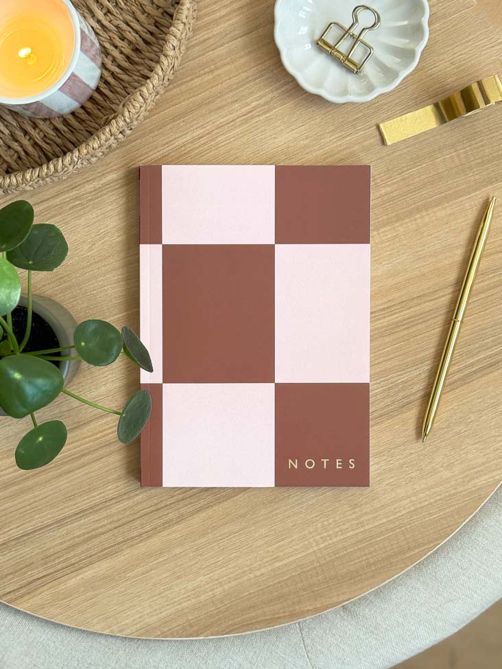 Checkered notebook with 'NOTES' embossed in gold foil on a wooden desk with stationery and desk accessories.