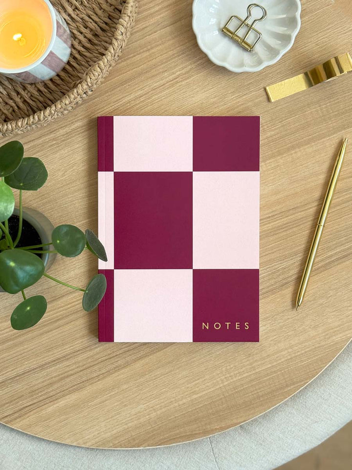pink and cherry red checkered A5 lined notebook with 'NOTES' in gold foil, on a wooden desk with decorative accessories.