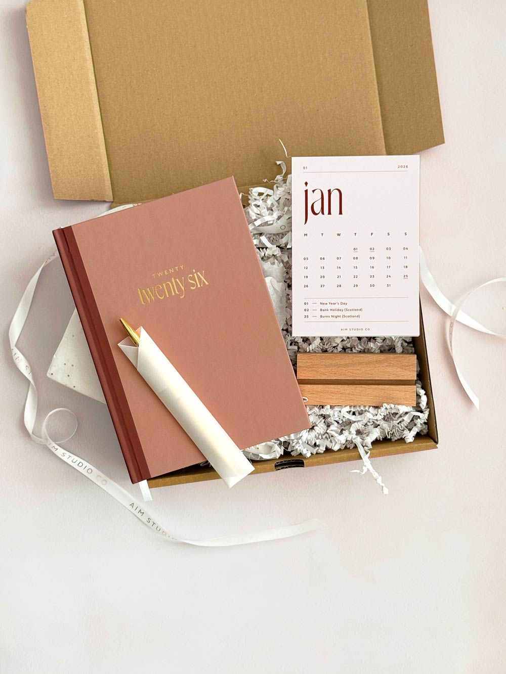 stationery gift set including a brown rust cover 2026 hardback diary with gold foiling, a slim gold pen in sleeve, and 2026 month view desk calendar cards with a wooden holder to display them. packaged in a box with decorative wrapping.