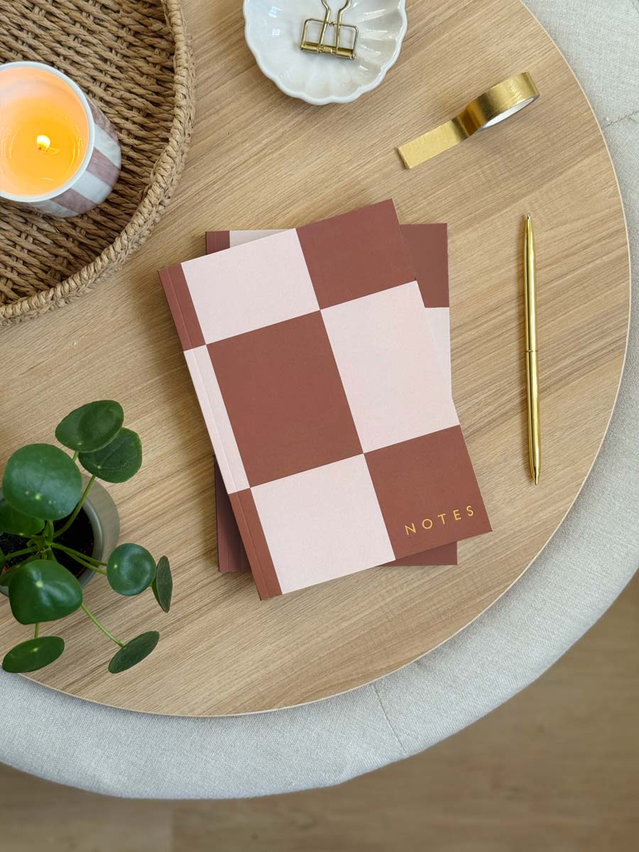 A5 rust brown Checkered notebook with 'NOTES' on a wooden table with a candle and plant.