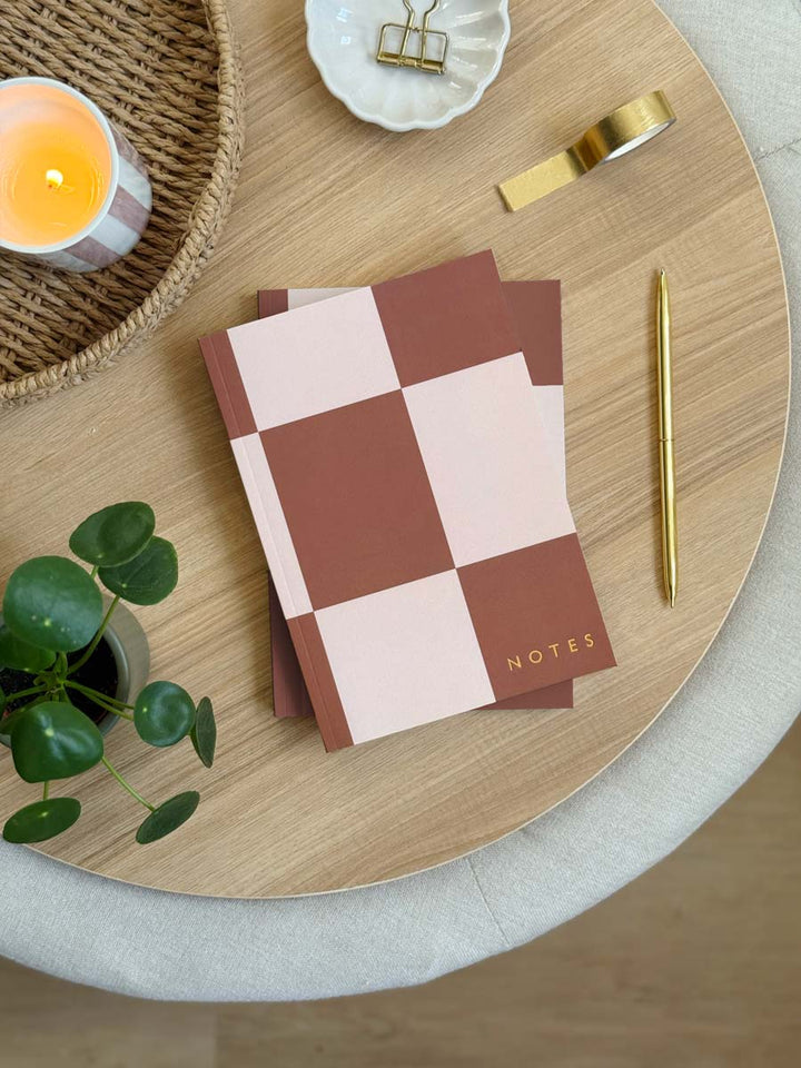 A5 rust brown Checkered notebook with 'NOTES' on a wooden table with a candle and plant.