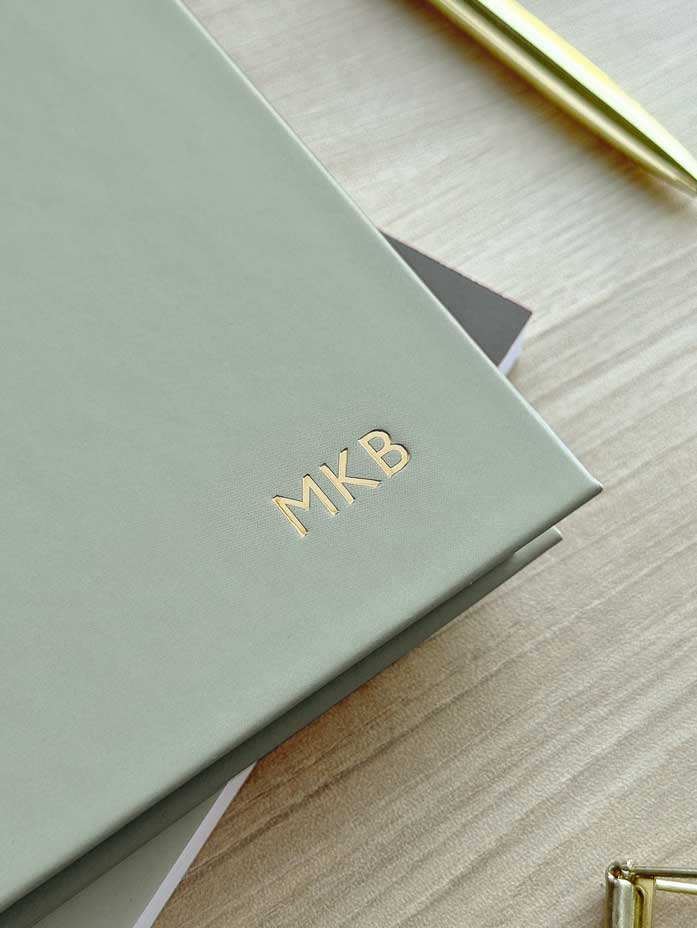 Sage green 2026 diary with 'MKB' initials embossed in gold foil and a gold pen on a wooden desk.