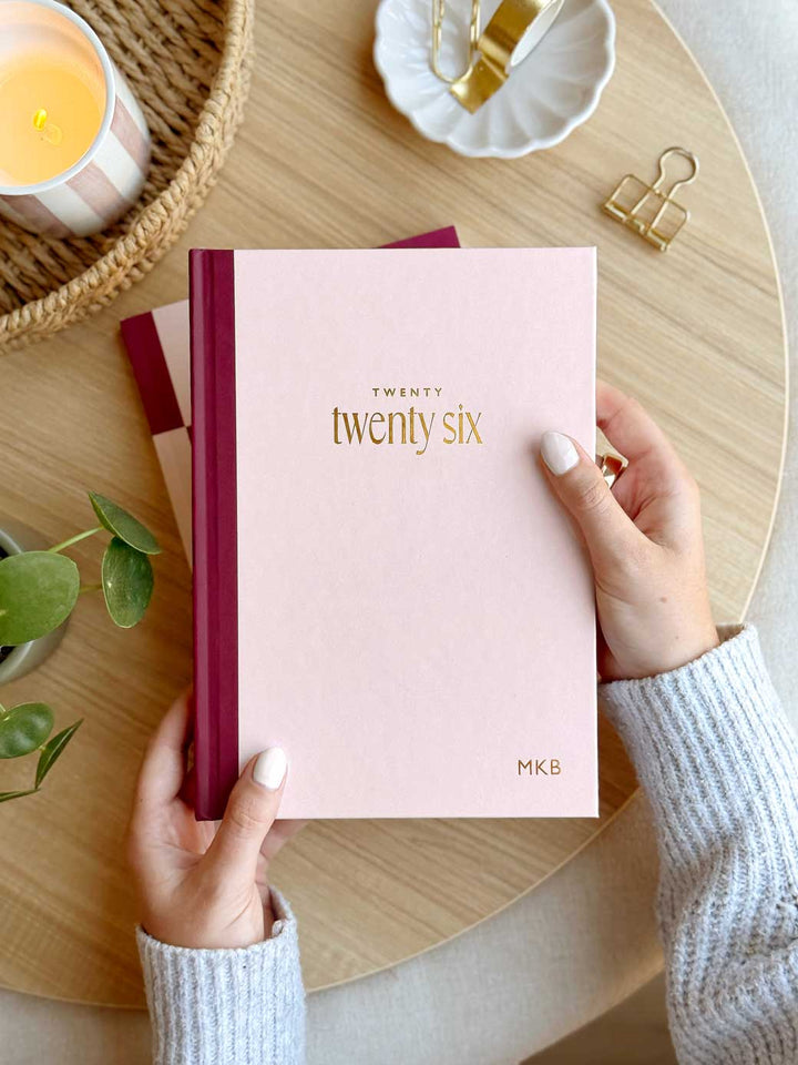 Woman holding a 2026 pink hardback diary with 'twenty twenty-six' on the cover in gold foil