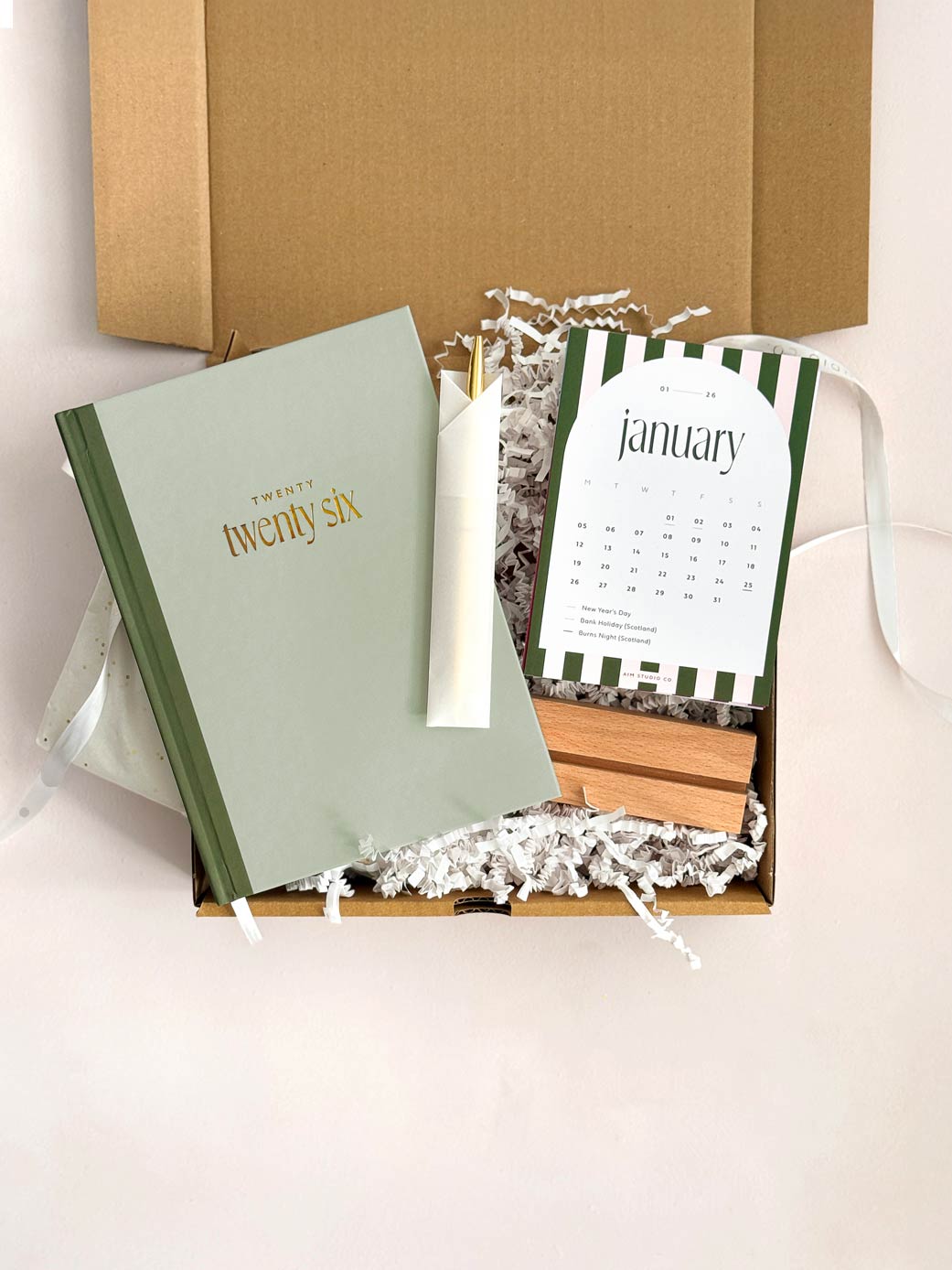 sage green hardback 2026 diary with gold pen next to a small desk calendar with wooden holder showing a stripe design for the month of January 2026. Packaged with decorative paper inside a cardboard gift box.