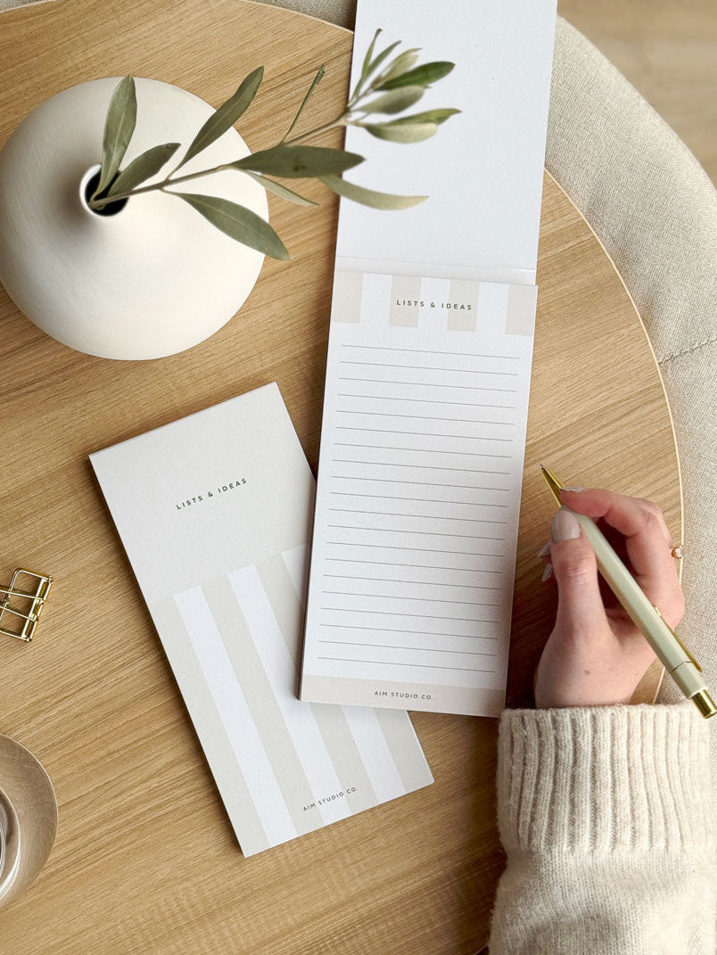 woman writing to do list on lined slim notepad by aim studio co featuring stripe design and surrounded by desk accessories and cute stationery