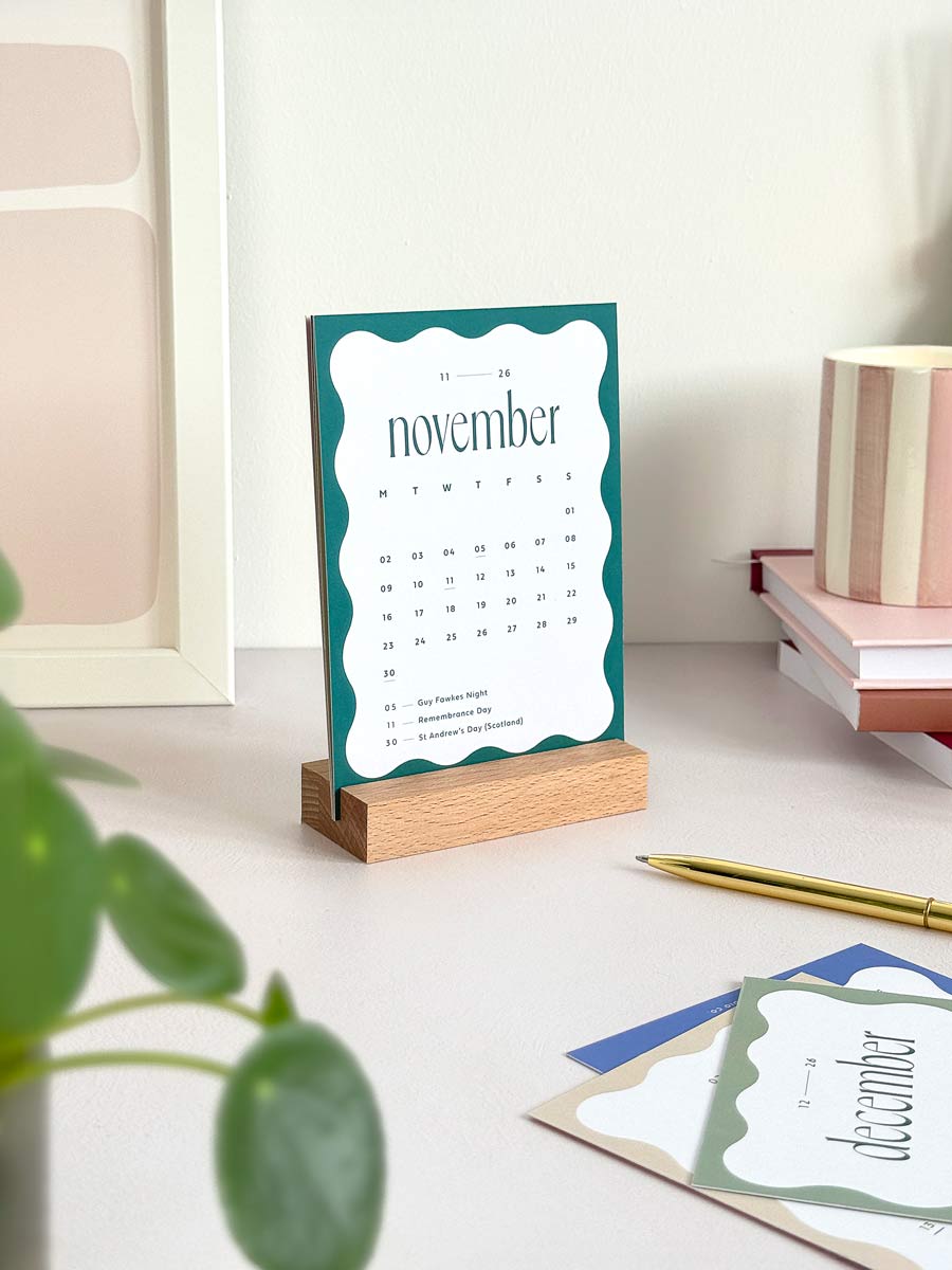 desk calendar with wavy border design in bold blue showing the month of november 2026 and UK bank holidays standing on an office desk with stationery and desk accessories.