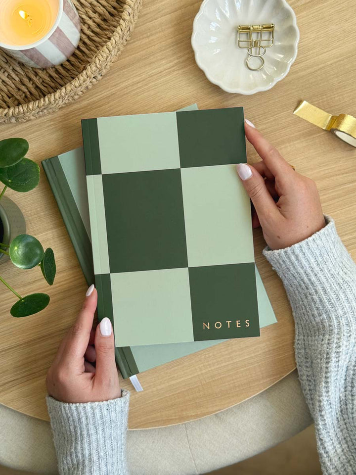 Person holding a green checkered notebook with 'NOTES' in gold foil on a wooden surface.