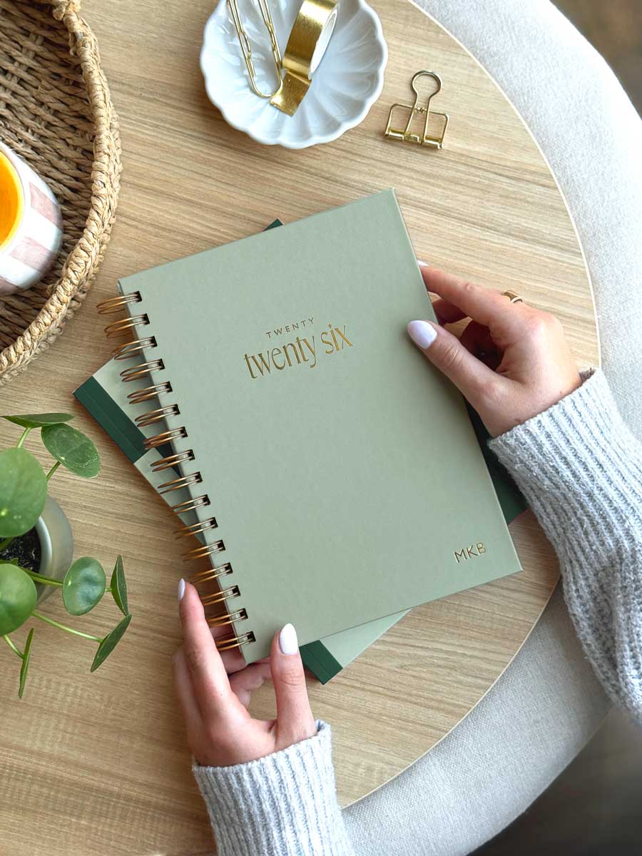 woman holding a sage green spiral hardback planner with gold foiled "2026" on the cover and personalised initials embossed in the corner of the diary cover.