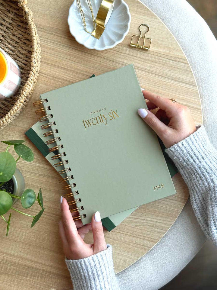 woman holding a sage green spiral hardback planner with gold foiled "2026" on the cover and personalised initials embossed in the corner of the diary cover.