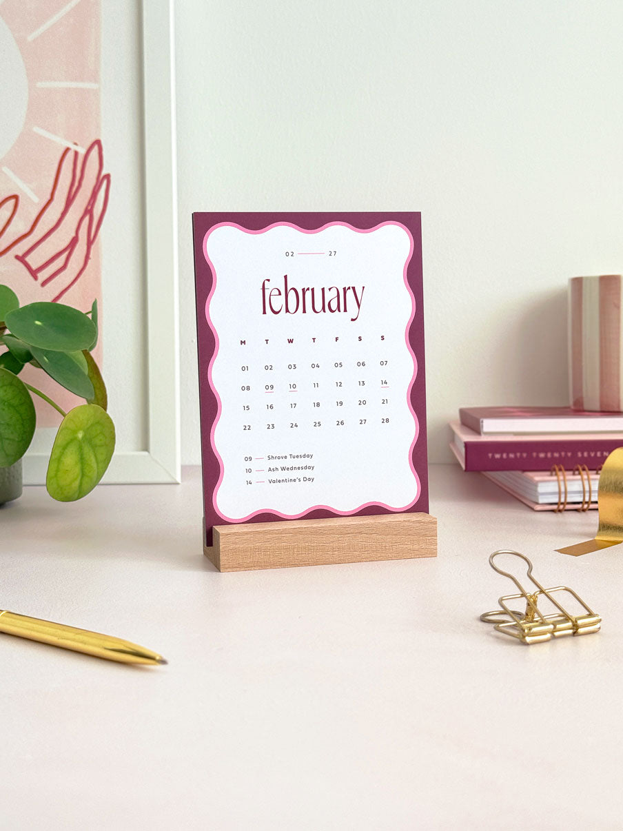 small desktop calendar card showing february 2027 held upright in a wooden stand. The A6 card features a deep burgundy and pink wavy border, a serif heading and a minimal date grid with UK occasions.