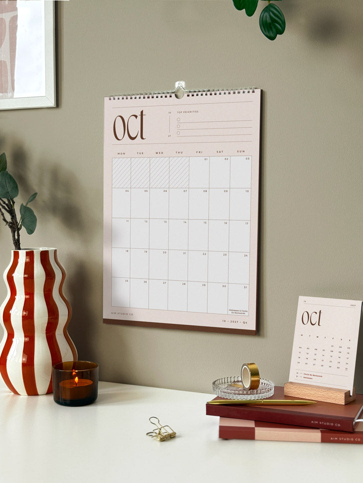 A spiral-bound 2027 wall calendar hanging on the wall above an office desk showing a neutral monthly page of october with planner grid. A smaller desktop version of the calendar sits on a stack of notebooks and diaries.