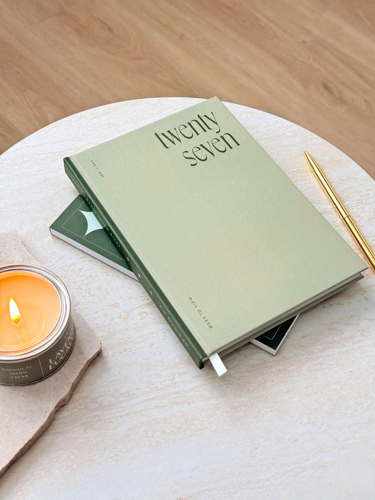 side view of hardback weekly planner with sage green cover and small text reading 'week to view' and 'jan - dec' on the cover. Styled on a white table with a gold pen and candle.