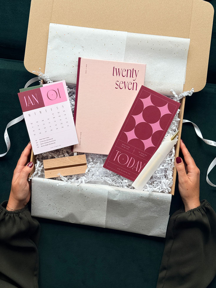 a gift box held by two hands showing a pink and burgundy stationery bundle with a hardback 2027 diary, geometric daily planner notepad, colour block desk calendar and a gold pen. The desk accessories are gift wrapped in white tissue paper inside a kraft box.