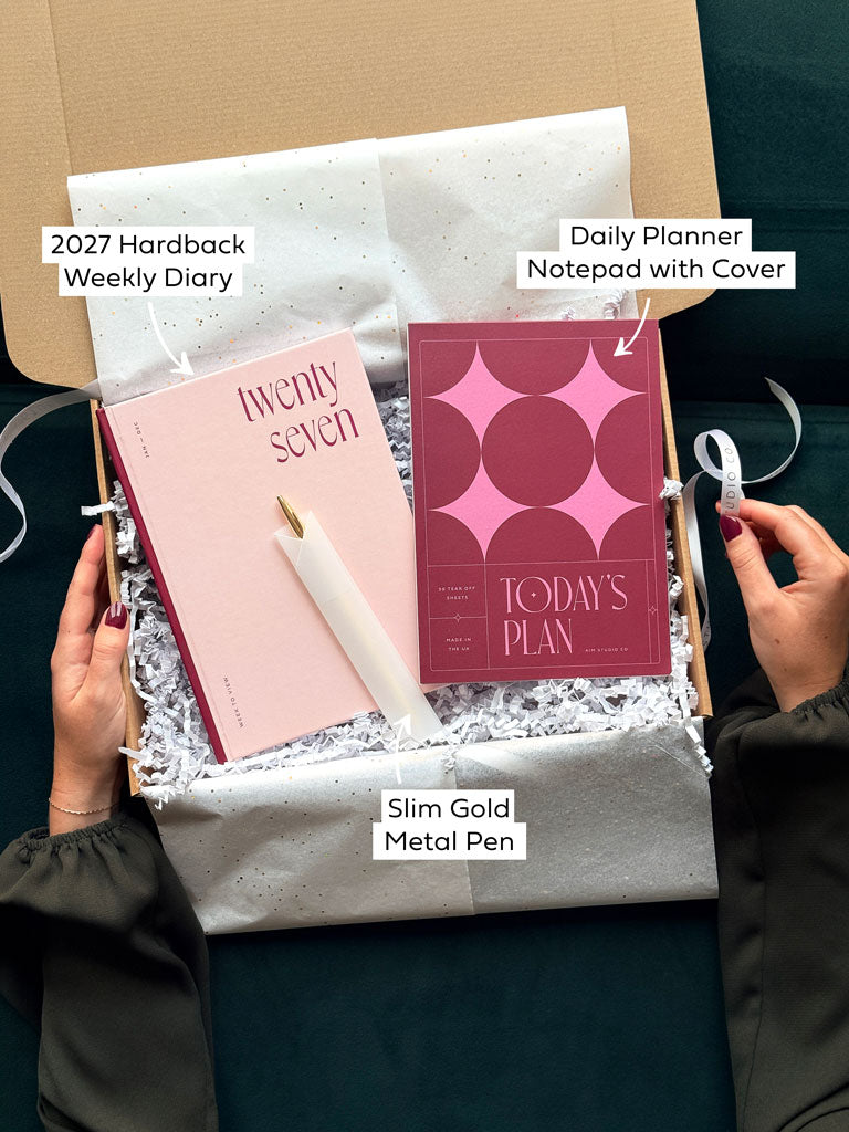 A stationery gift box containing a 2027 hardback weekly diary in light pink, a daily planner notepad with a pink cover, and a slim gold metal pen, arranged on white tissue paper. Two hands are holding the box, and text labels identify each item.
