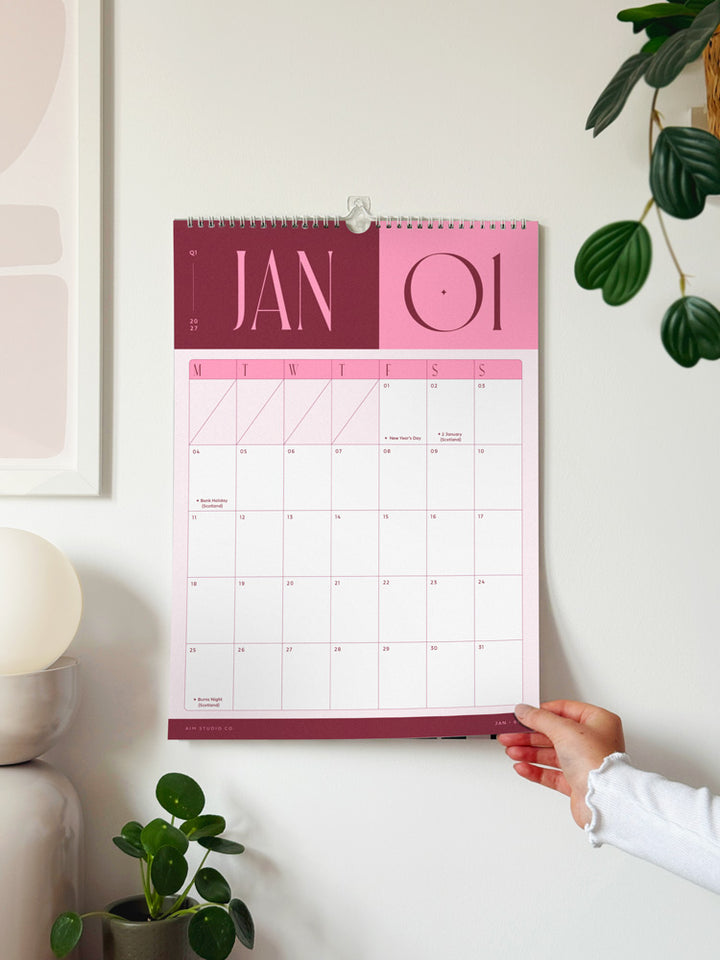 A pink and burgundy bold wall calendar is hanging on a white wall showing the monht of January 2027. The calendar features a clean grid layout and is styled in a bright, modern room with a plant, lamp, and framed art.