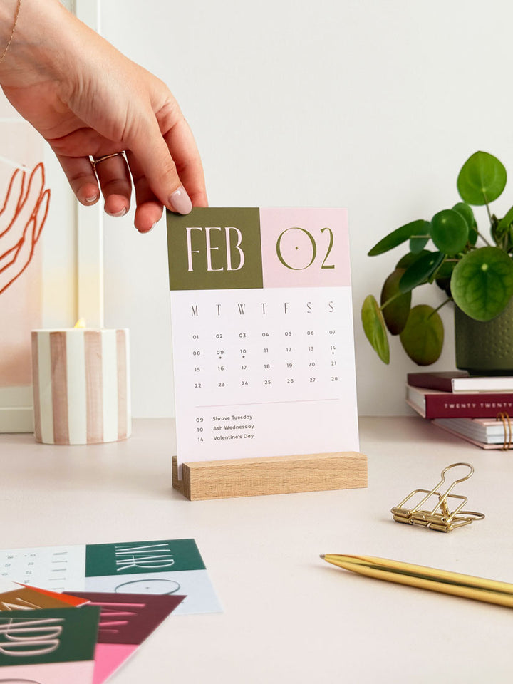 Minimalist desk decor surrounding a 2027 refillable monthly desk calendar. a woman is changing the olive green and pink desk calendar card showing february 2027 held in a wooden stand.