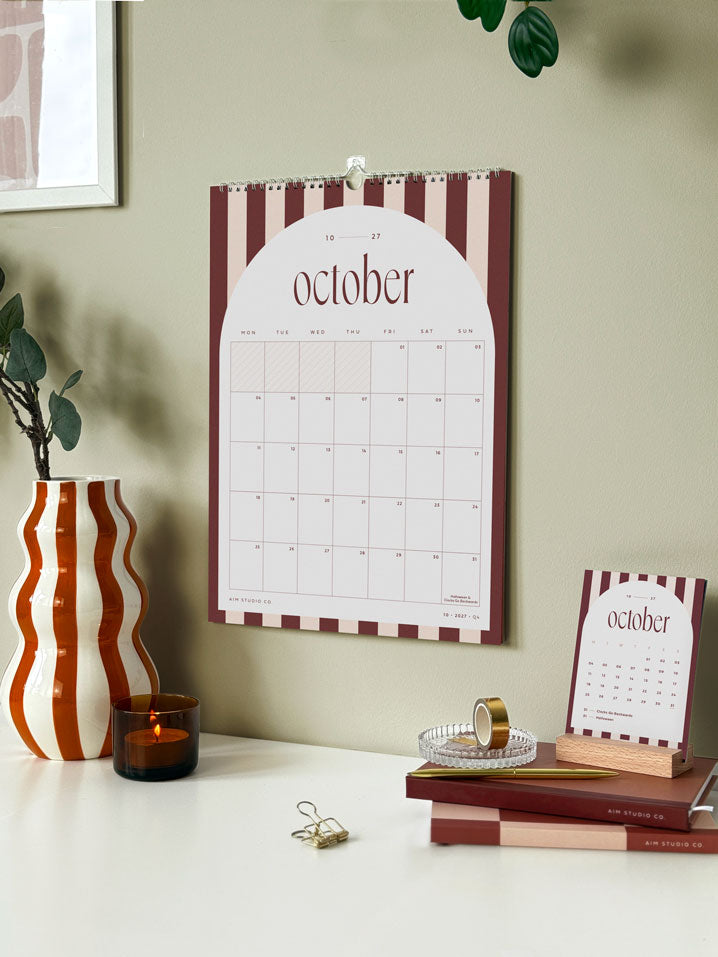 monthly wall calendar showing brown stripe design for october hanging on an office wall above a desk. matching 2027 desk calendar cards held in a wooden stand are displayed beneath the calendar on a stack of diaries and notebooks.