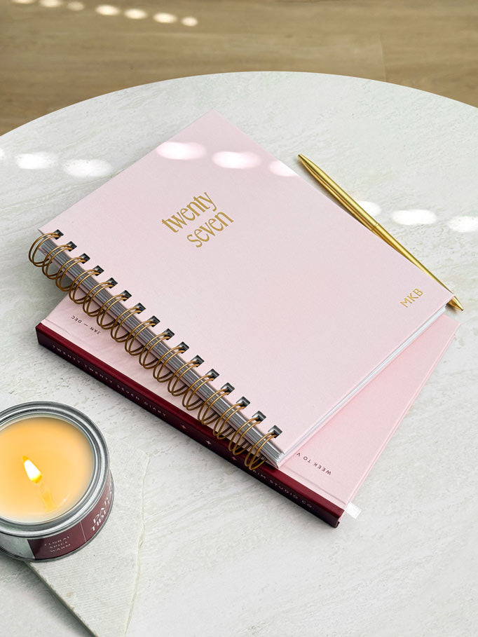 A pink spiral-bound weekly planner with gold foiled lettering reading &