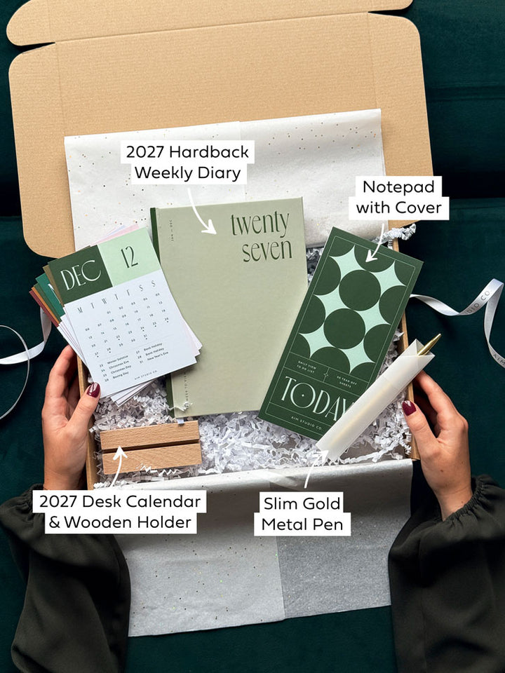A stationery gift box with text highlighting each item, containing a sage green themed set of desk accessories, planner and notepad with a pen.