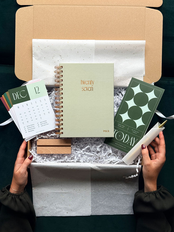 A styled flat-lay of a stationery gift box opened by two hands. Inside the kraft cardboard box with white tissue paper and shredded filler are a pale sage green spiral notebook with gold foil text reading &