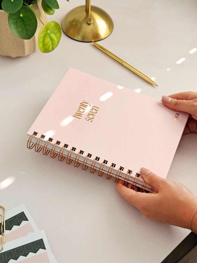 woman placing a pale pink 2027 personalised diary on an office desk. Surrounded by planner stickers sheets, a gold pen, lamp and a potted plant.