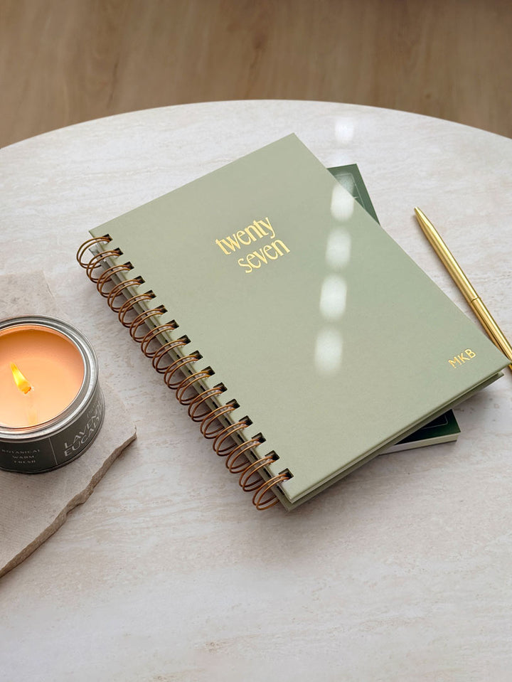 2027 diary with sage green hardback cover and gold foiled details. The planner has metallic spiral binding and lies on a white marble desk.
