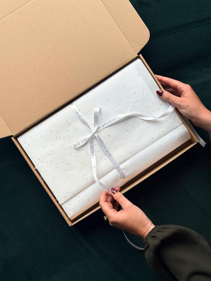 An open cardboard box with aesthetic white tissue paper with gold flecks, tied with an AIM Studio Co braned ribbon. A womans hands are unwrapping the gift bundle to reveal the stationery gift set inside.