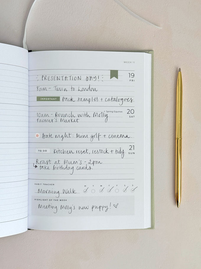An open weekly planner notebook showing handwritten notes and a calendar layout, photographed on a light beige surface with a gold pen beside it.