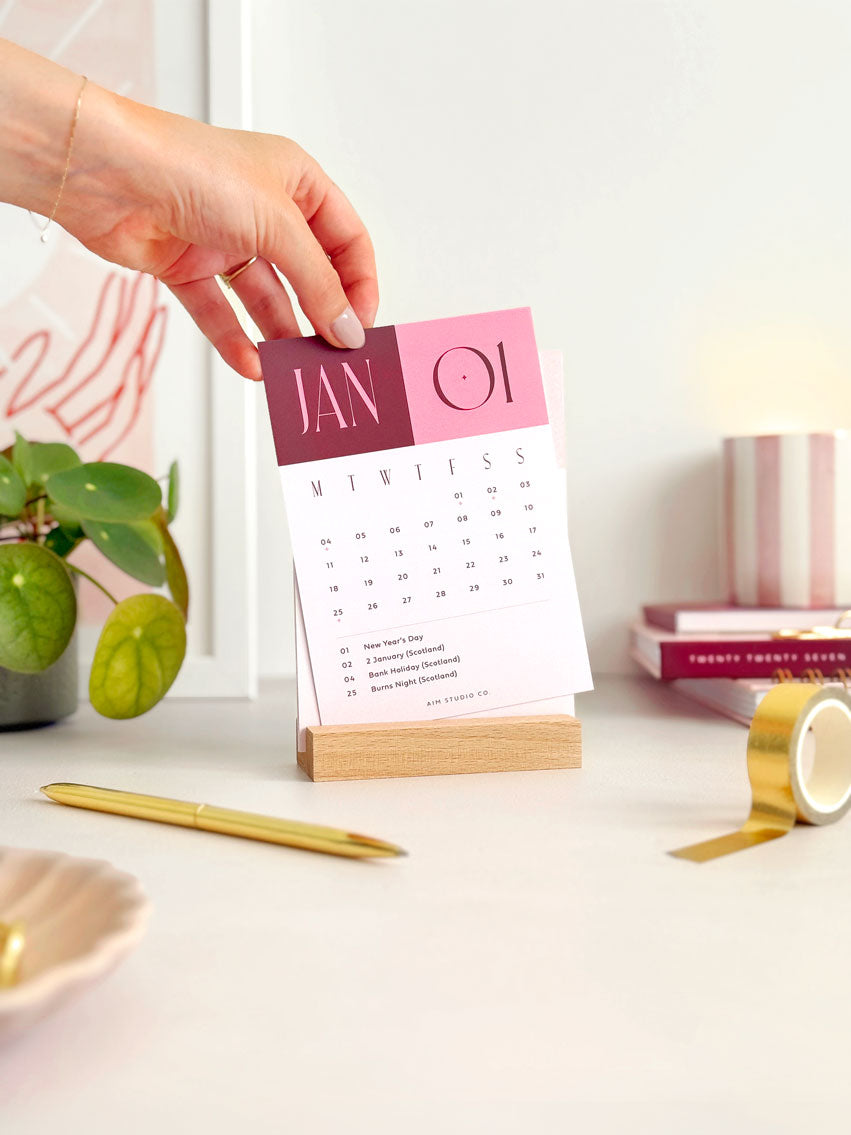 a6 sized monthly desk calendar card showing January 2027 sitting in a wooden block stand on an office desk.
