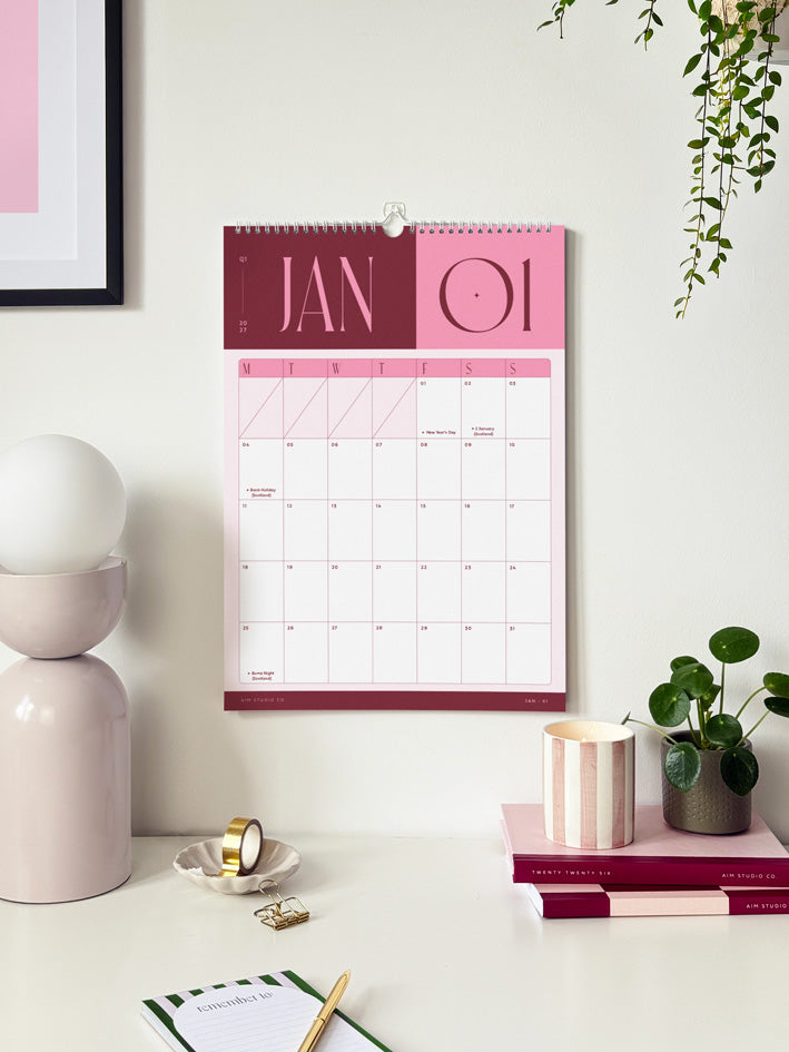 A large a3-sized pink and burgundy wall planner showing January 2027 monthly calendar page hanging on an office wall with desk accessories and stationery beneath it.
