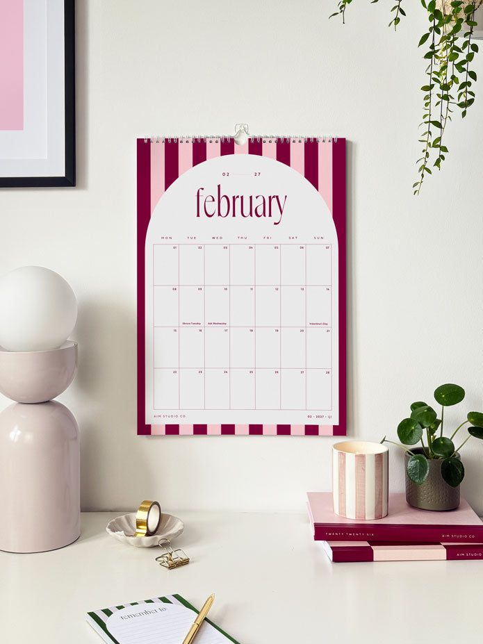 Striped wall calendar showing the month of february 2027 with pink candy striped design on an office wall above a styled desk.