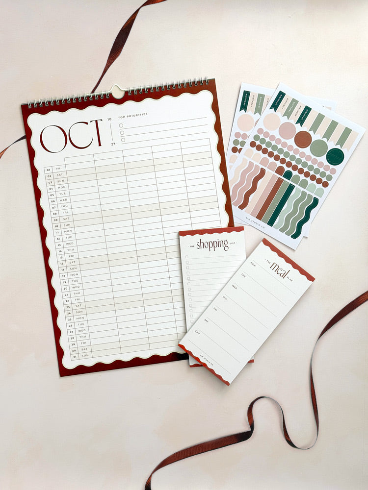 The Family Organiser Set in rust: flat lay of a large A3 2027 family wall planner calendar with stickers and a matching wavy weekly meal planner and shopping list in rust brown.