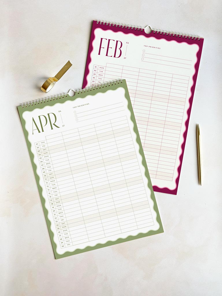 two large a3-sized family calendars on a desk - one shows february 2027 in deep pink and the other shows april 2027 in sage green. Both monthly planner pages have a bold wavy border and vertical planner grid with 5 columns.