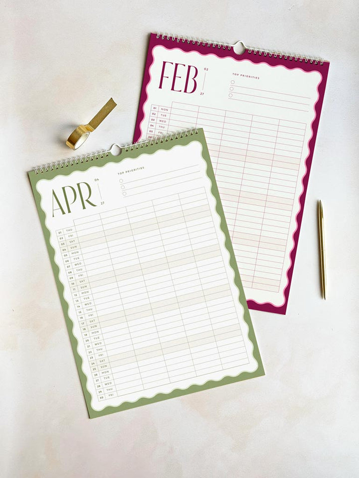 two large a3-sized family calendars on a desk - one shows february 2027 in deep pink and the other shows april 2027 in sage green. Both monthly planner pages have a bold wavy border and vertical planner grid with 5 columns.