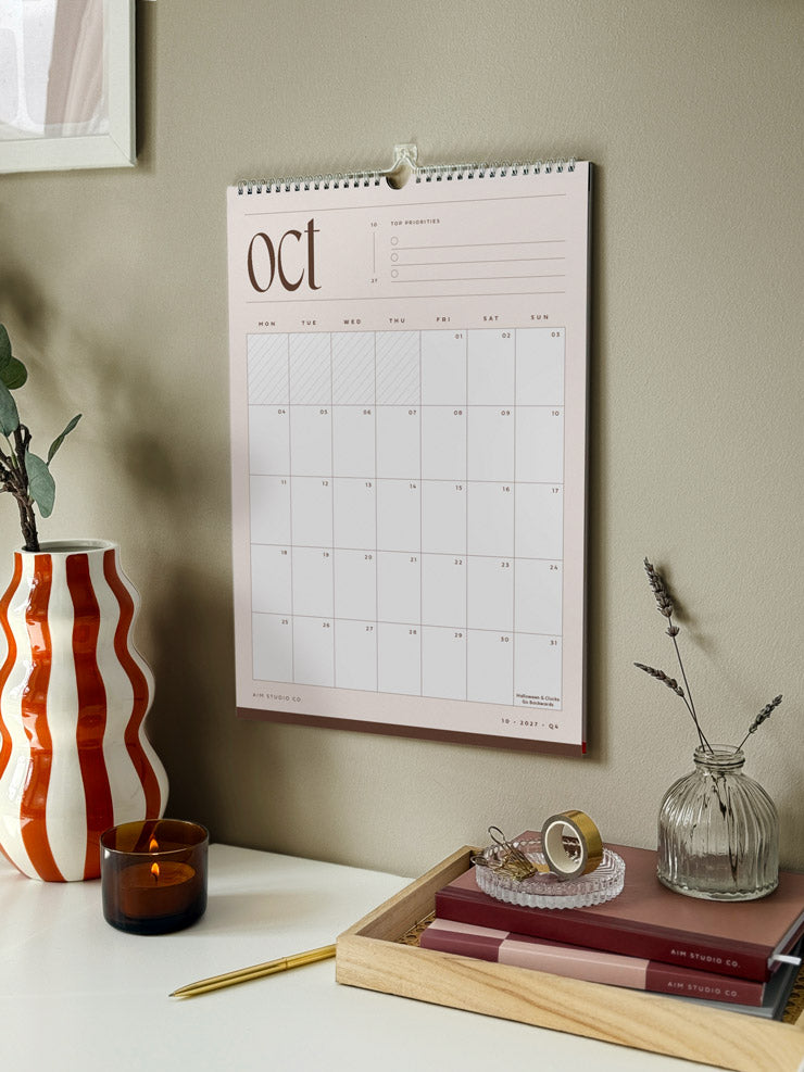 A monthly calendar hanging on a sage green wall, styled in a home office setting with desk accessories. The wall planner shows the month of october 2027 in neutral beige and brown colours.
