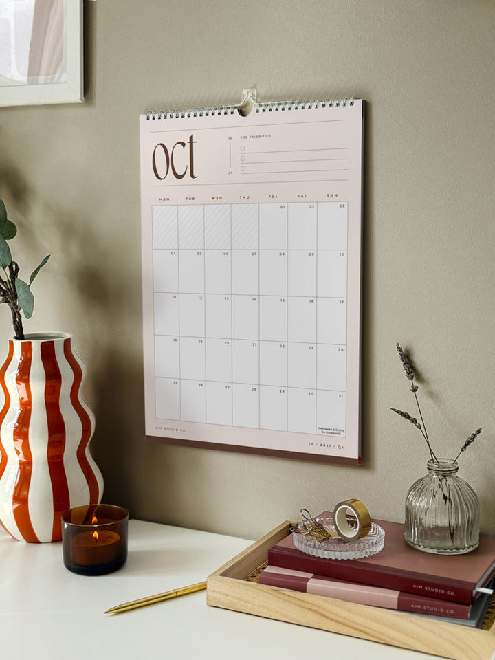 A monthly calendar hanging on a sage green wall, styled in a home office setting with desk accessories. The wall planner shows the month of october 2027 in neutral beige and brown colours.