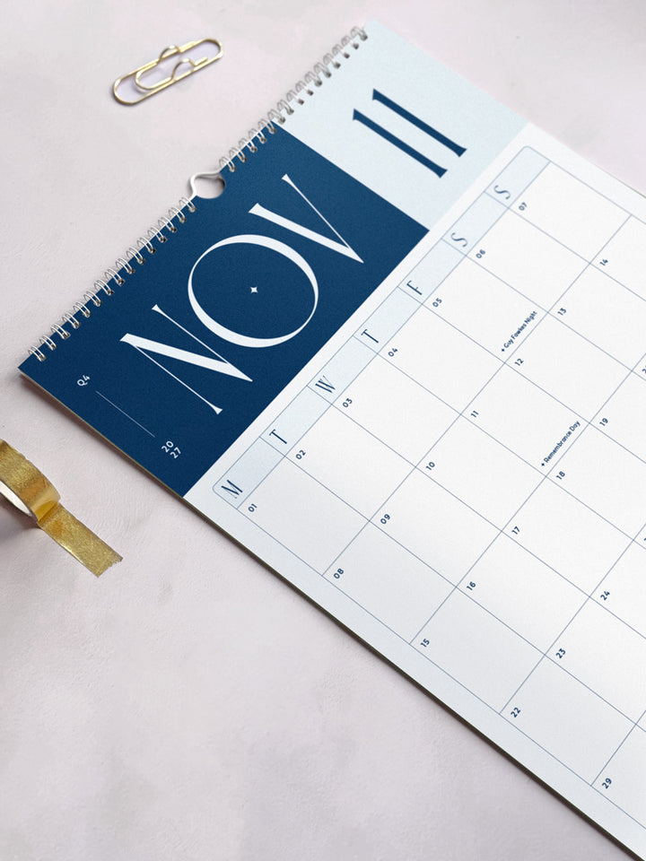 A spiral-bound 2027 monthly wall planner showing November calendar page with a navy blue header and white grid layout with monday start.