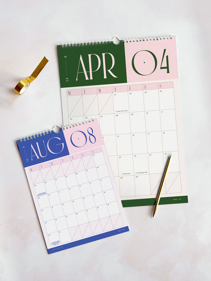 Two wall calendars laid on a light surface: one for April and one for August, each with a large date header in green and pink and a monthly grid below. Features white spiral binding with a hanging loop.