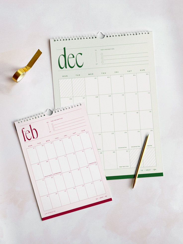 a small a4 calendar and a large a3 wall calendar laid on a desk surface. One shows december 2027 in sage green and the other shows a pink and red february month page. Both calendars have white wire binding and feature a monthly planner grid with UK holidays marked.