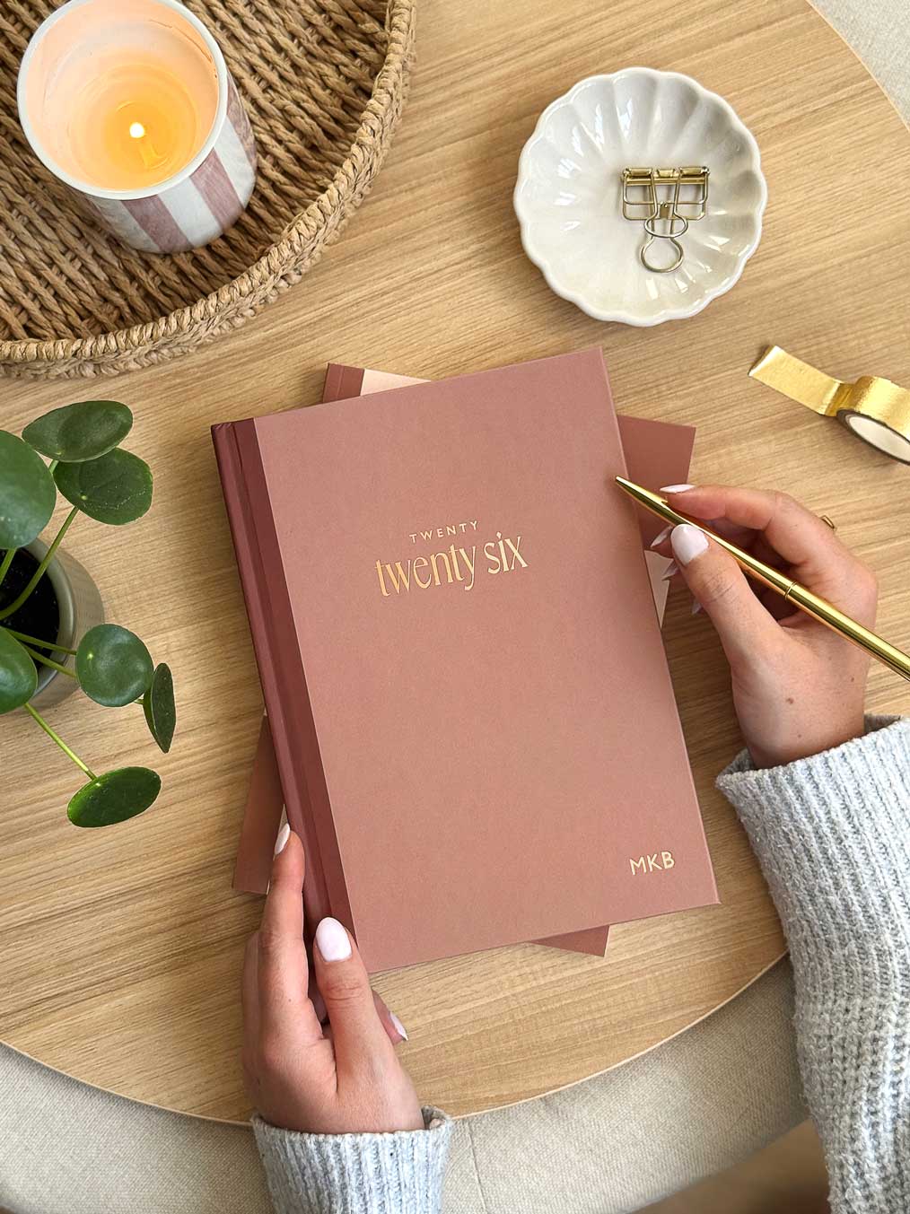 Person holding a rust coloured 2026 diary with 'Twenty Six' font in gold foil on a wooden desk with a plant and desk accessories 