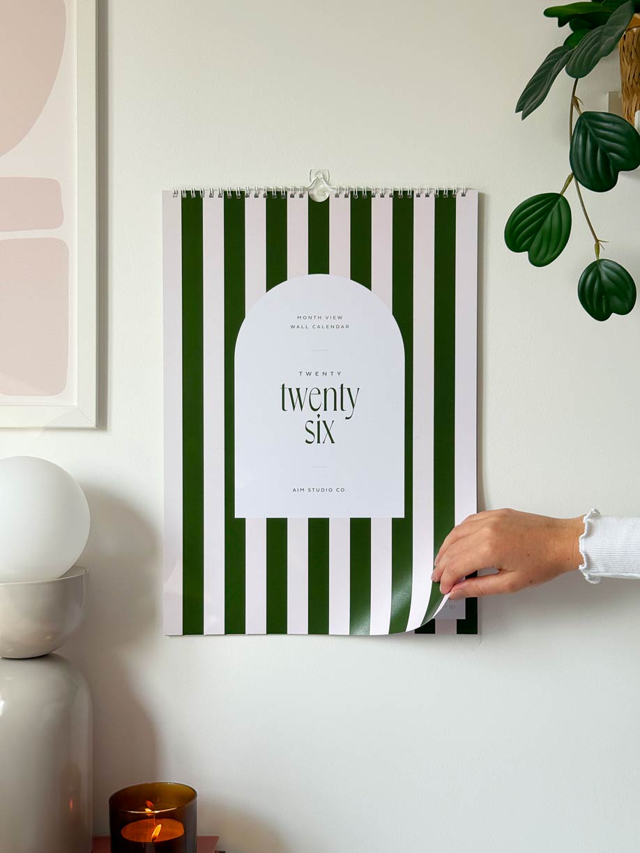 green and pale pink striped wall planner in large a3 size hanging on office wall with woman turning cover page to reveal monthly calendars inside. 