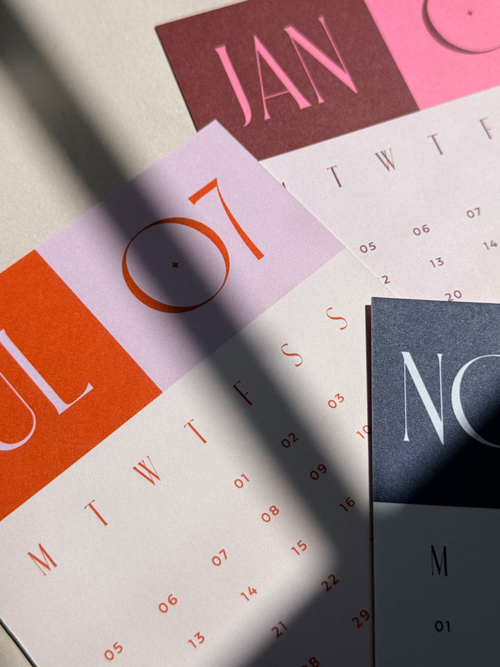 Close up of naturally uncoated smooth paper texture on 350gsm printed 2027 calendar cards in assorted colourful designs. The monthly cards are scatted on a desktop surface with bright sunlight and cast shadows.