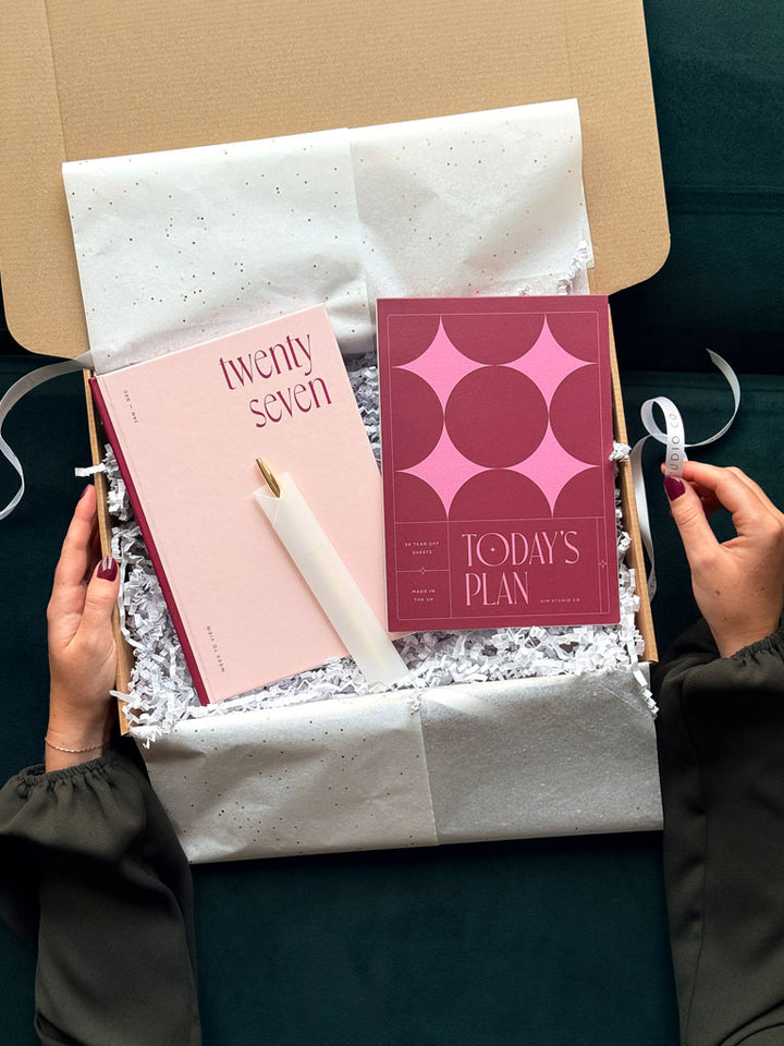 a stationery gift box containing a pink hardback 2027 diary with a deep burgandy and pink daily planner notepad. A slim gold pen lies ontop of the planners and hands are holding either side of the gift box.