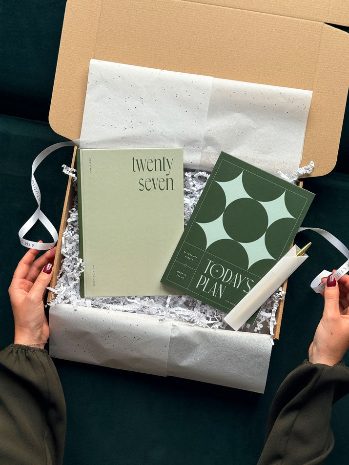 flat lay of a stationery gift box containing a sage green 2027 weekly diary, an a5 daily planner notepad with deep green cover and a slim gold pen, presented inside a gift wrapped bundle box.