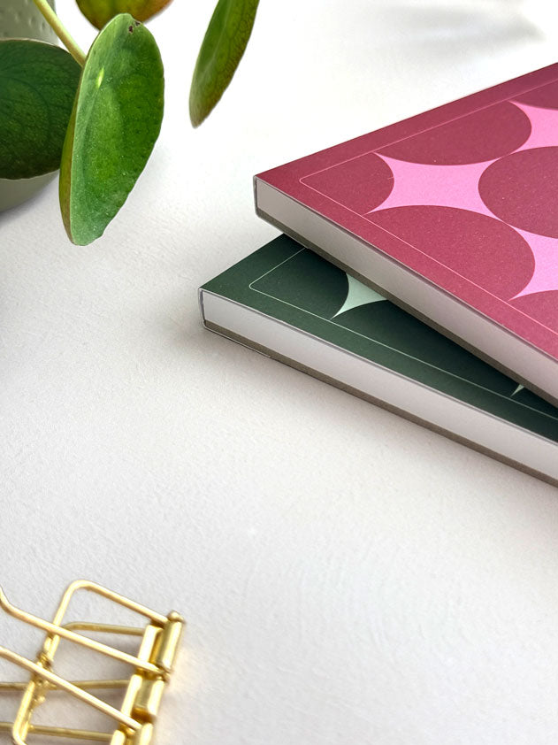 two geometric notepads with covers stacked ontop of each other on a light desk surface. The design features a bold retro star in a geometric, grid-line layout - one in a burgundy and pink colour way and one in deep green with pale green stars.