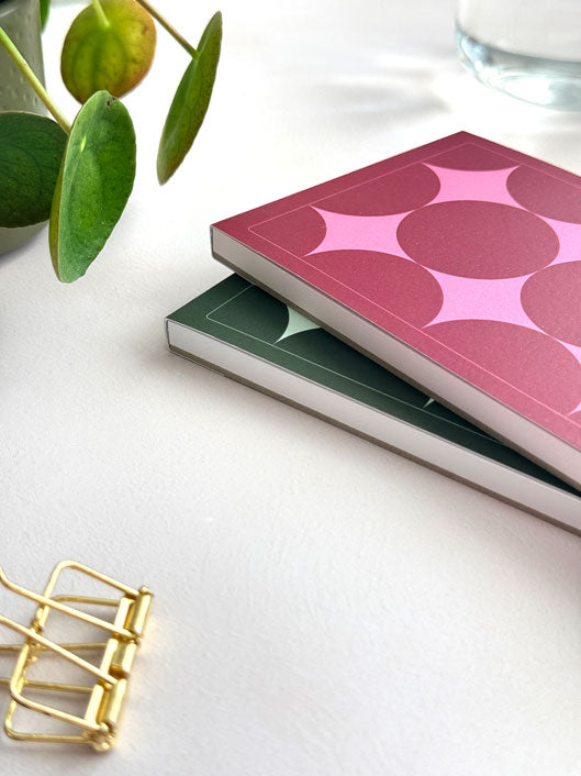 Side view of two stacked daily notepads with star design on the cover. The top notepad featured a deep red and pink colour way, and the bottom one shows the design in a deep green version. Surrounded by a plant and gold clip on a desk.