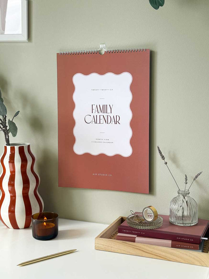 hanging wall calendar showing cover of 2026 family planner on a sage green wall with bold home accessories.